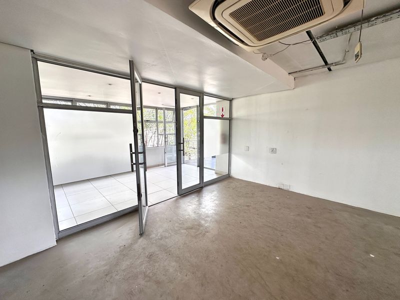 An empty office space featuring exposed ceiling infrastructure, a ceiling-mounted air conditioning unit, and aluminium-framed glass doors leading to an adjacent patio area.