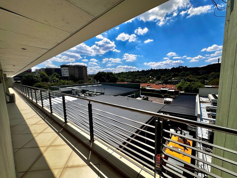 A spacious covered balcony featuring tiled flooring and a modern stainless steel balustrade, offering an elevated view of the surrounding office park and landscape.