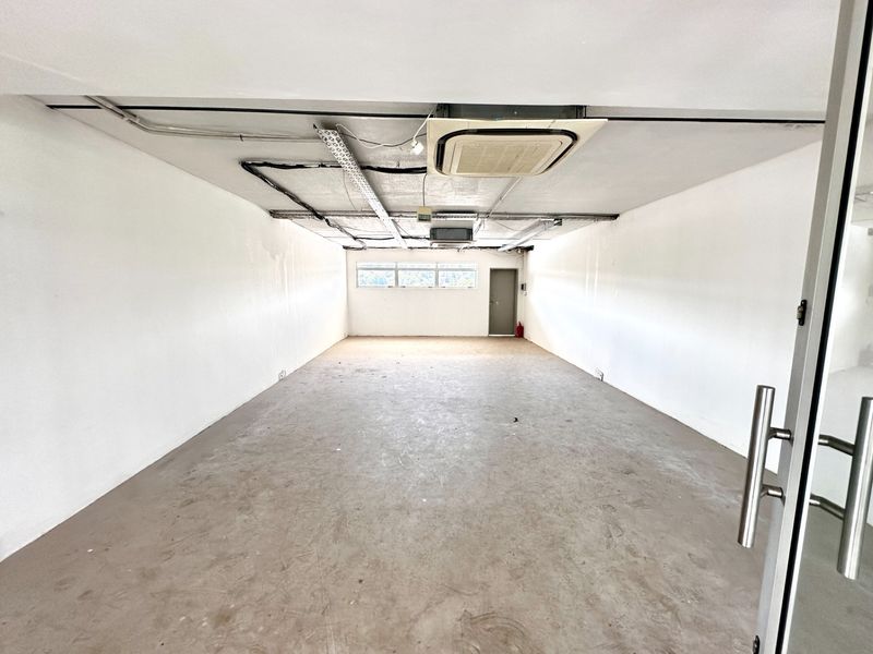 An empty, open-plan office space featuring exposed ceiling infrastructure, concrete flooring, and a ceiling-mounted air conditioning unit.