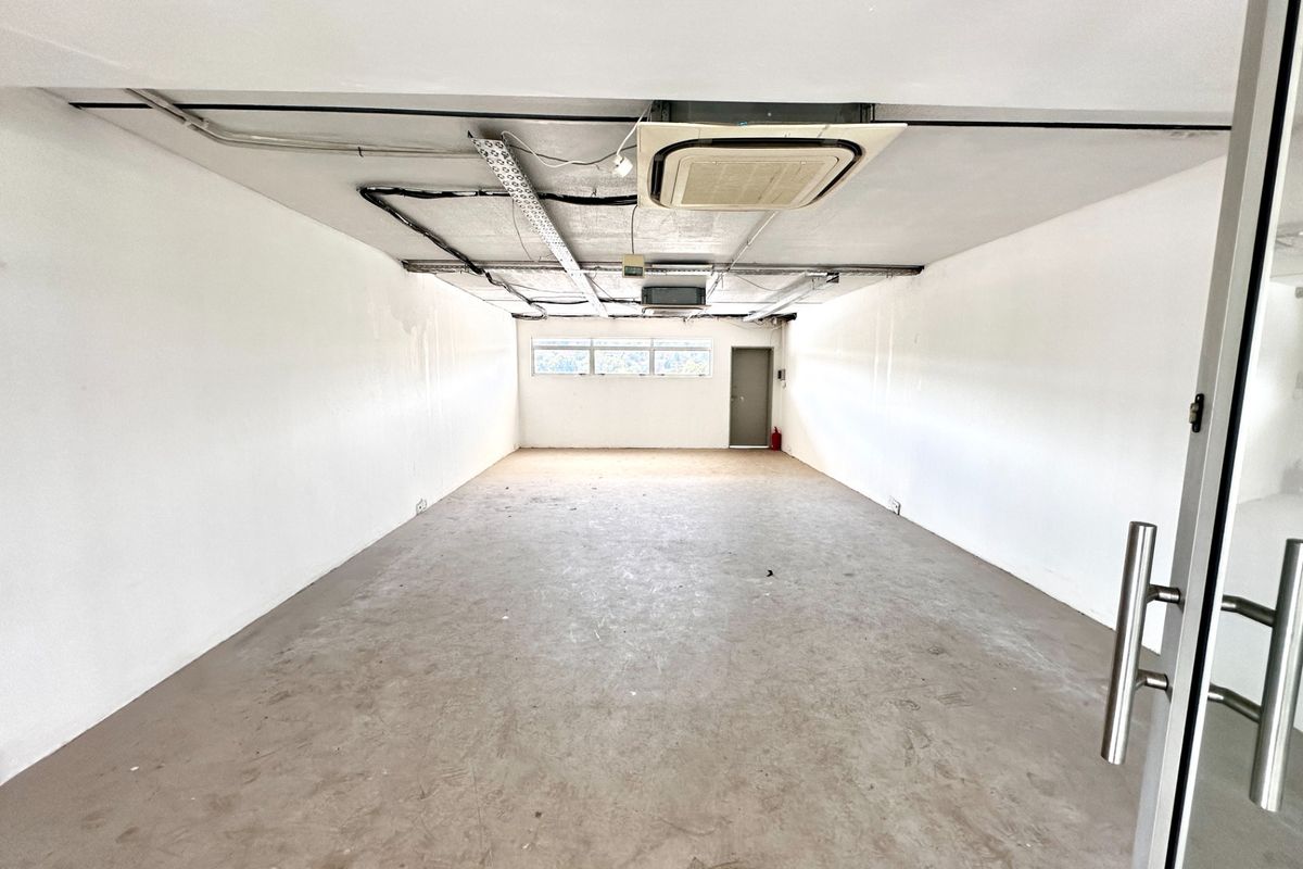 An empty, open-plan office space featuring exposed ceiling infrastructure, concrete flooring, and a ceiling-mounted air conditioning unit.