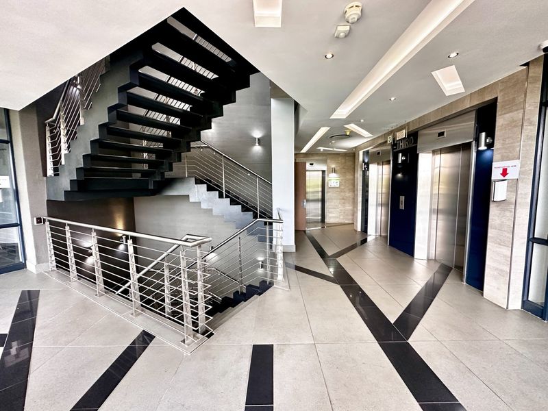 Modern office building lobby featuring a prominent staircase with stainless steel railings, tiled flooring, and multiple elevator access points.