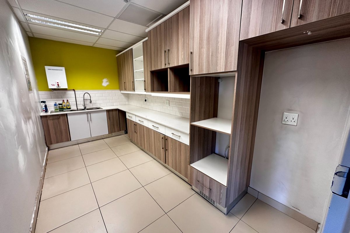 A functional office kitchenette featuring wood-look laminate cabinetry, white subway tile backsplash, stainless steel sink, and tiled flooring in a clean, open layout.