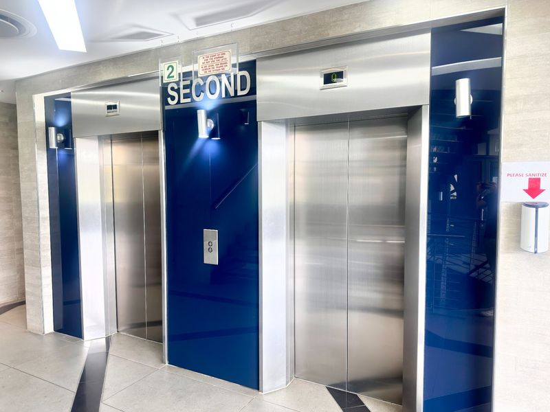 A modern office building elevator lobby featuring stainless steel lift doors, polished tiled flooring, and clean, contemporary wall finishes.
