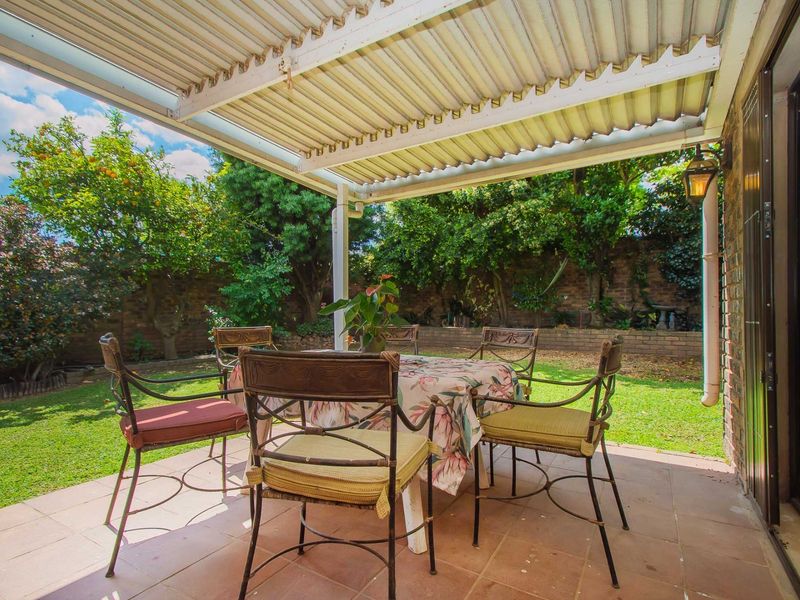 A covered patio area featuring tiled flooring and a corrugated roof, overlooking a private garden with established trees and a brick boundary wall.