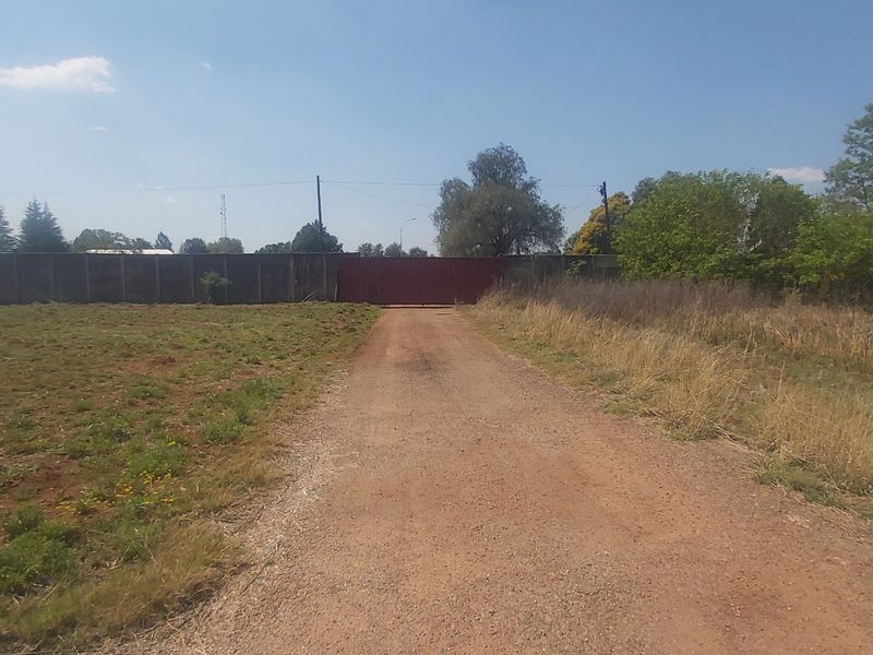 A view of a vacant residential plot featuring a dirt driveway leading to a gated entrance and perimeter walling.