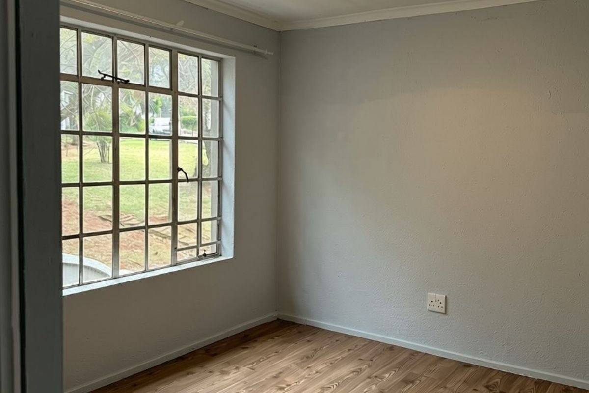 A bright, empty bedroom featuring wood-look laminate flooring, a large grid-pattern steel window, and neutral wall finishes.