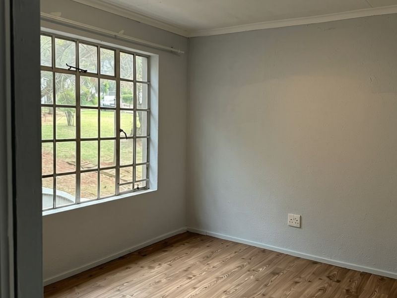 A bright, empty bedroom featuring wood-look laminate flooring, a large grid-pattern steel window, and neutral wall finishes.