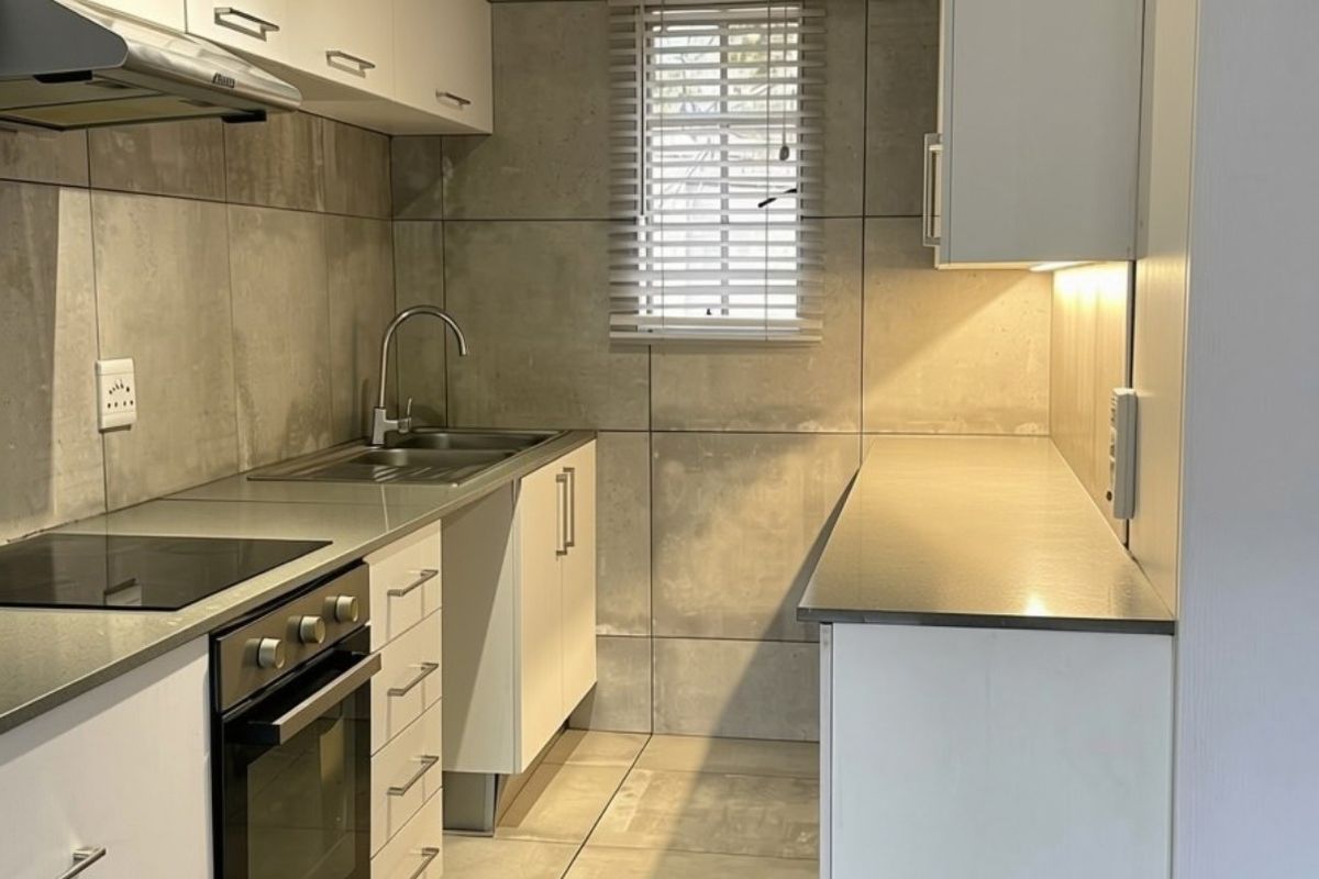 A modern, clean kitchen featuring white cabinetry, stainless steel appliances, tiled walls, and ample counter space, illuminated by under-cabinet lighting.