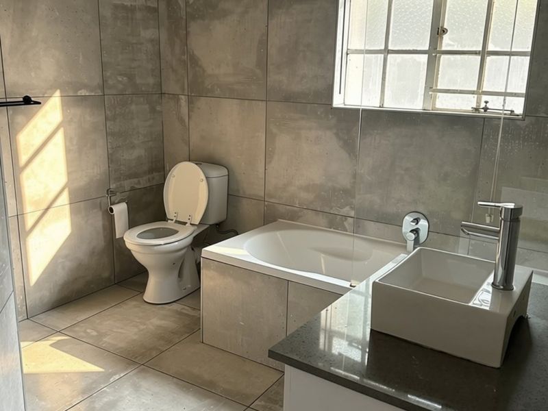Modern bathroom featuring large format grey tiles, a built-in bathtub, a vessel sink on a stone vanity, and a toilet.
