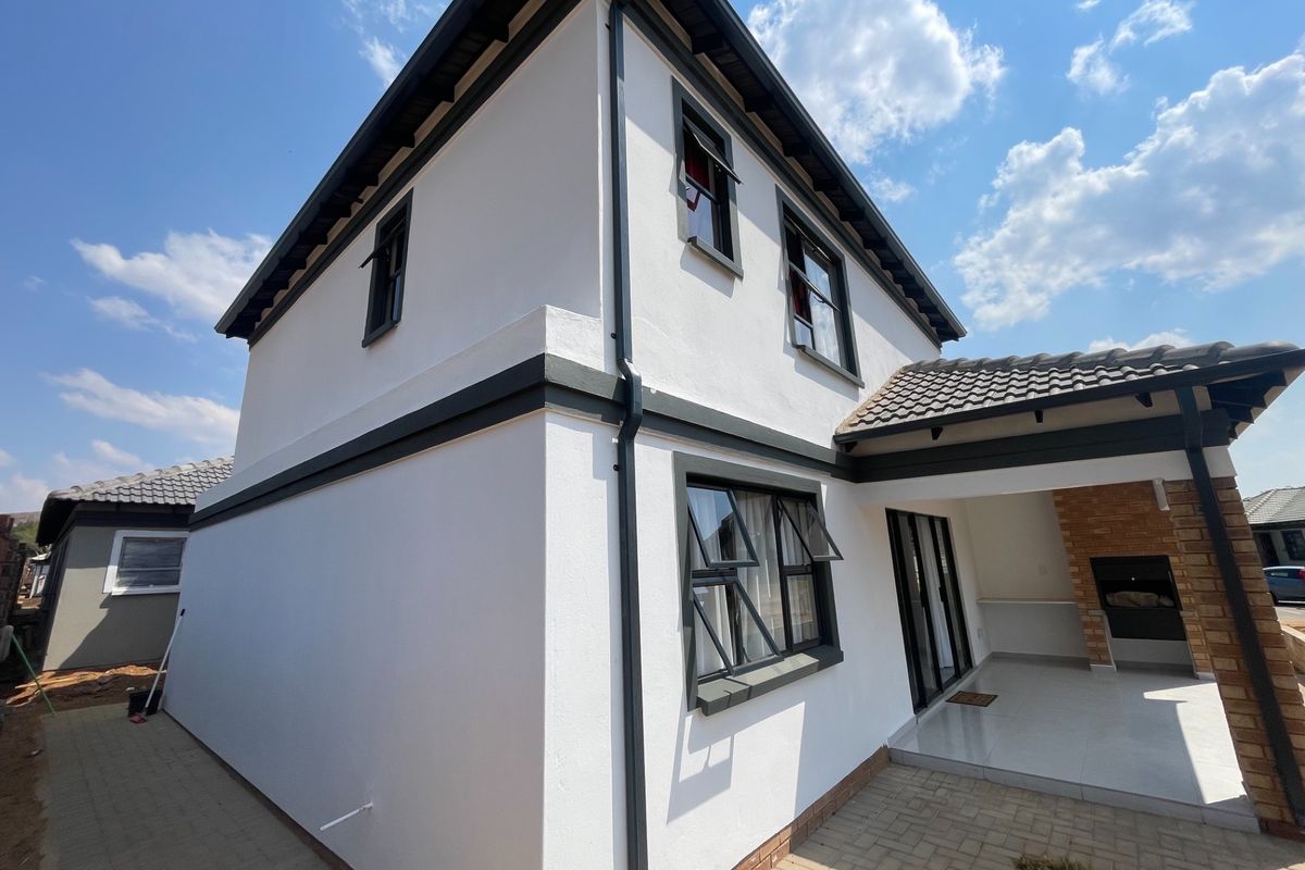 A modern double-storey home exterior featuring a paved patio area with a built-in braai, clean white render, and dark aluminium window frames.