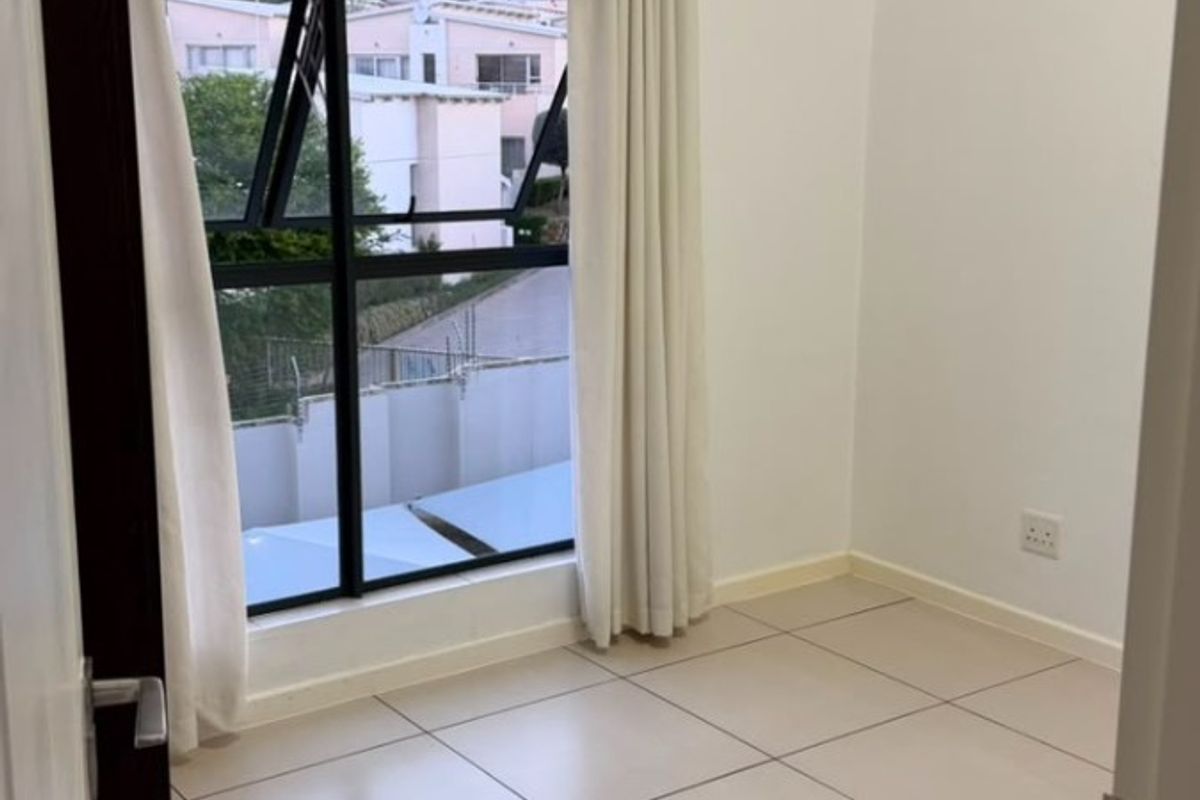 A bright, empty bedroom featuring neutral tiled flooring, clean white walls, and a large aluminium-framed window with natural light.