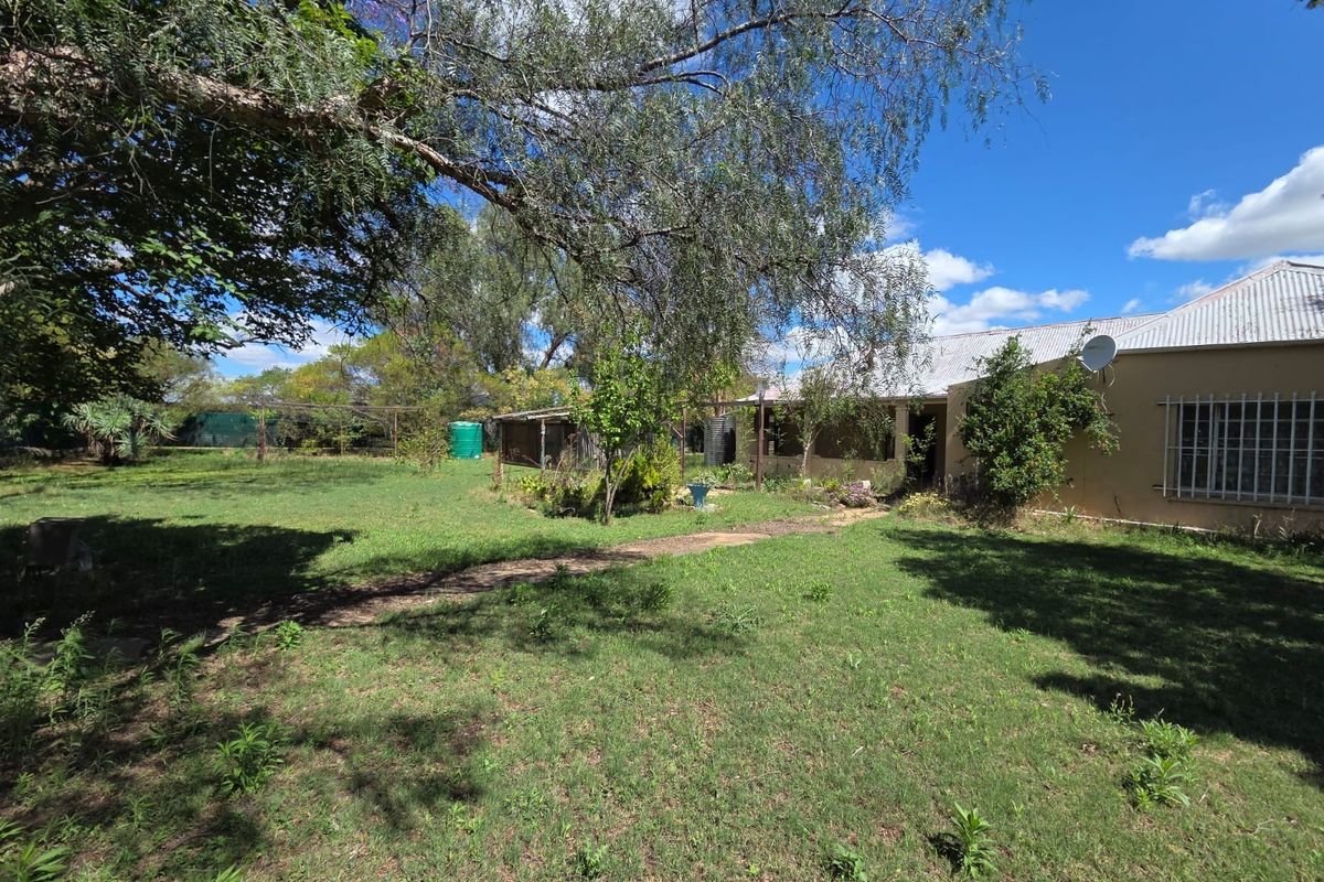 A view of the rear exterior of the property featuring a large lawn, mature trees, and a corrugated metal roof structure.