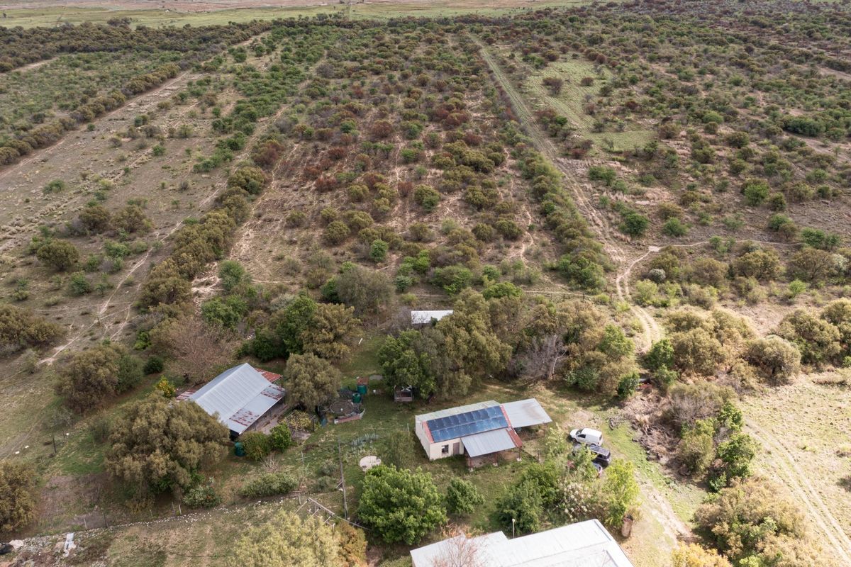 Aerial view of a small holding property featuring multiple structures with metal roofing, solar panel installations, and extensive surrounding natural vegetation and land.