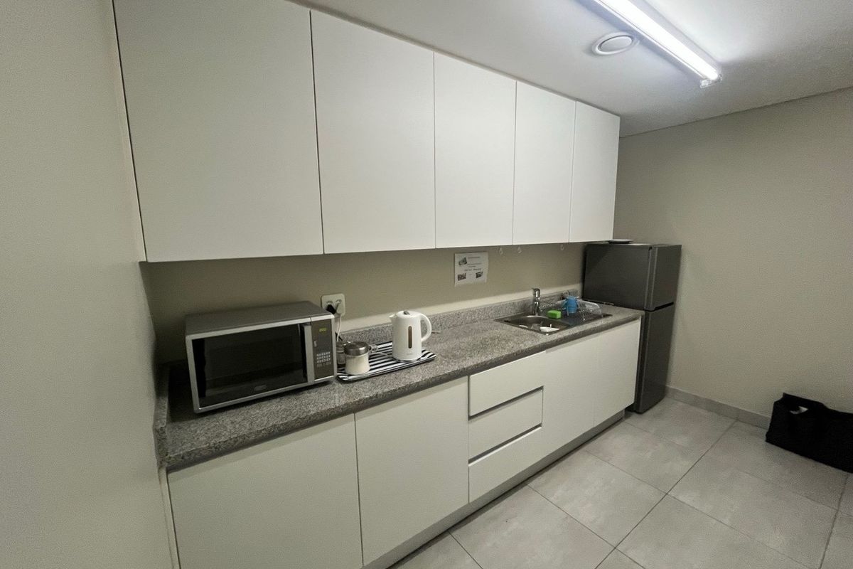 A functional kitchenette featuring white cabinetry, stone countertops, a stainless steel sink, and tiled flooring, suitable for office or industrial utility use.