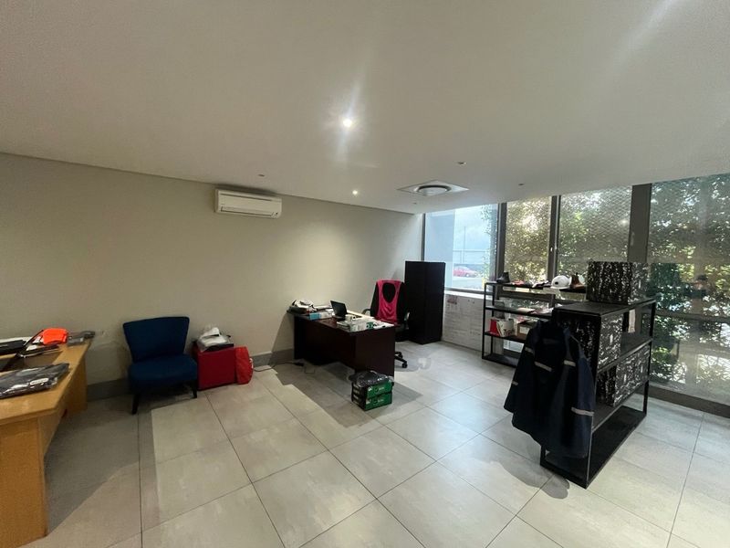 A functional office space featuring tiled flooring, recessed lighting, a wall-mounted air conditioning unit, and large windows providing natural light.