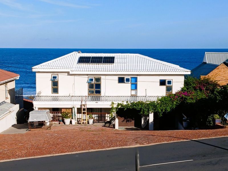 Front elevation of a coastal house featuring a metal roof with solar panels, a paved driveway, and a covered ground-level patio area.