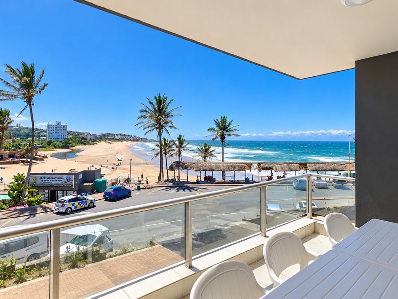 A spacious balcony featuring a modern glass balustrade and tiled flooring, offering expansive, unobstructed views of the beach and coastline.