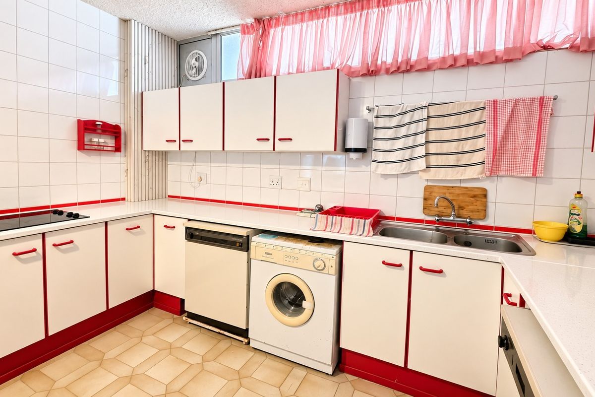 A functional kitchen featuring white cabinetry with red trim, double basin sink, integrated hob, and tiled splashbacks, offering a dated but practical layout.