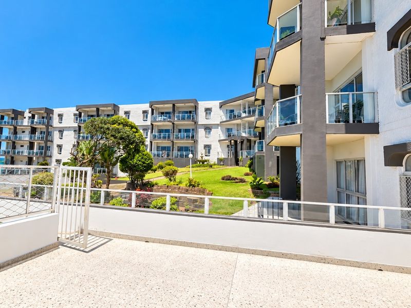 A spacious rear balcony with terrazzo flooring and glass balustrades, overlooking a well-maintained communal garden and the apartment complex exterior.