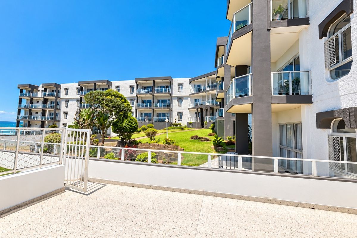 A spacious rear balcony with terrazzo flooring and glass balustrades, overlooking a well-maintained communal garden and the apartment complex exterior.