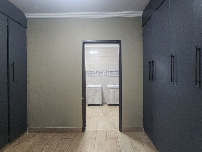 A spacious dressing area featuring extensive built-in cupboards with modern finishes, leading directly into a tiled bathroom with dual vanity units.