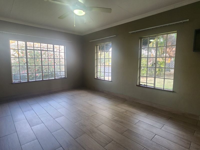 A spacious, empty living room featuring modern wood-look floor tiles, a ceiling fan, and large windows providing ample natural light.
