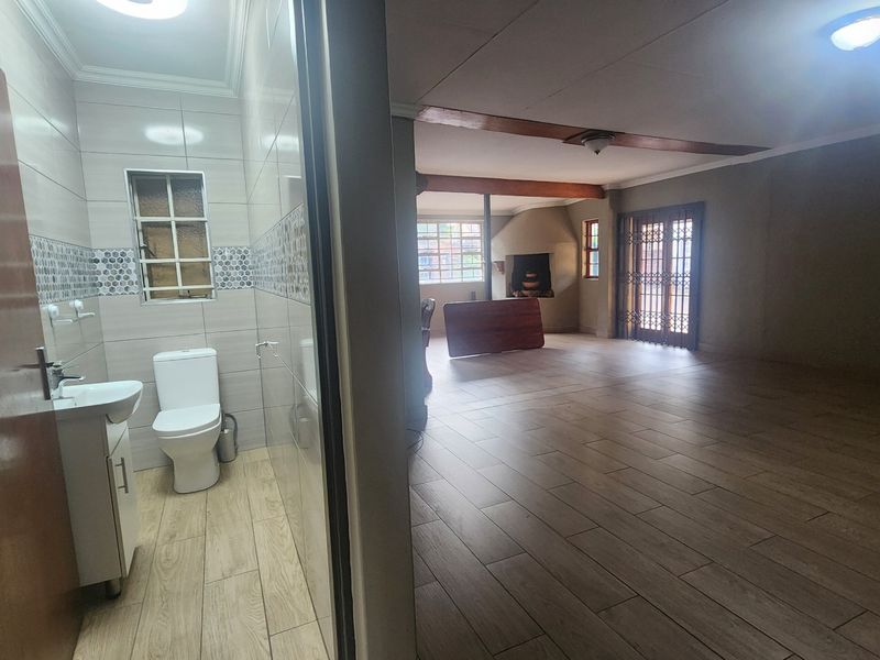 An open-plan living area featuring wood-look tiled flooring, exposed ceiling beams, a built-in fireplace, and an adjacent modern bathroom with a vanity and toilet.