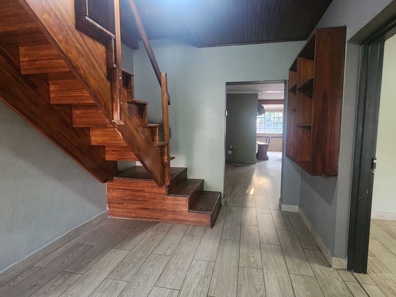 A spacious hallway featuring a prominent wooden staircase, built-in wall shelving, modern tiled flooring, and timber ceiling paneling.