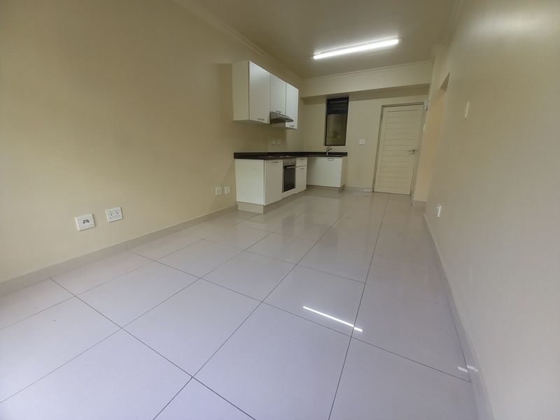 An open-plan living and kitchen area featuring tiled flooring, built-in cabinetry, and integrated kitchen appliances in a clean, vacant space.