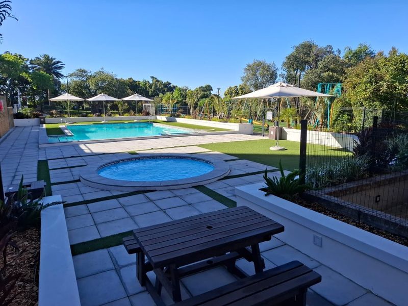 A well-maintained communal pool area featuring a swimming pool, a circular splash pool, paved patio space, and artificial lawn areas.