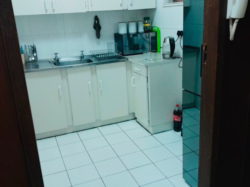 A compact kitchen featuring white built-in cabinetry, a stainless steel sink, and tiled flooring, viewed through a doorway.