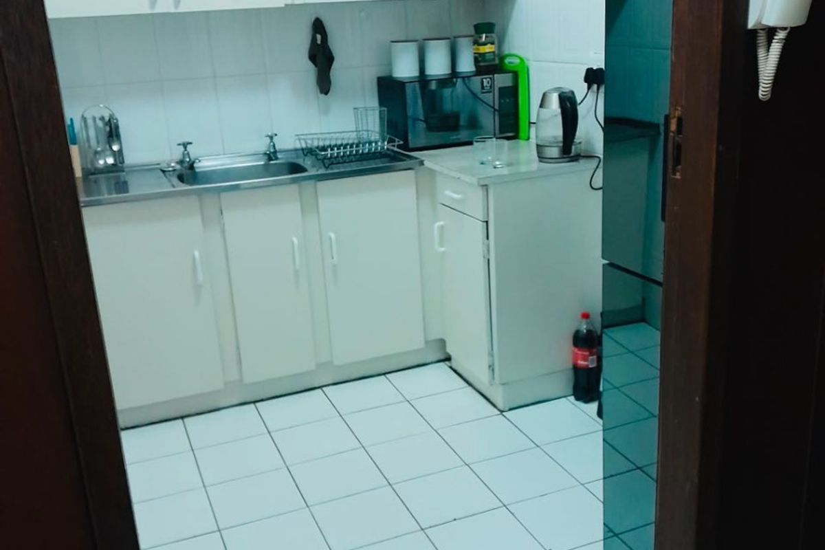 A compact kitchen featuring white built-in cabinetry, a stainless steel sink, and tiled flooring, viewed through a doorway.