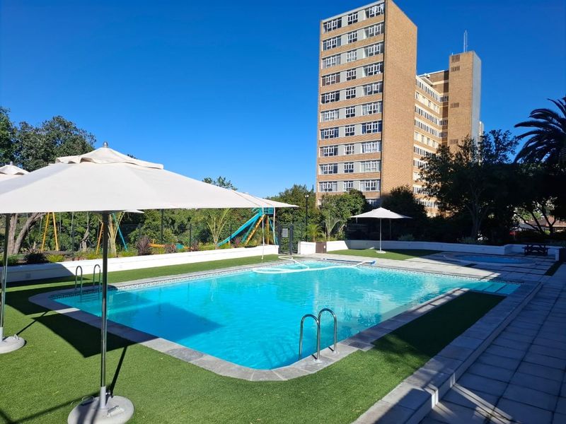 A well-maintained communal swimming pool area featuring artificial turf surrounds, paved decking, and perimeter fencing, set within an apartment complex.