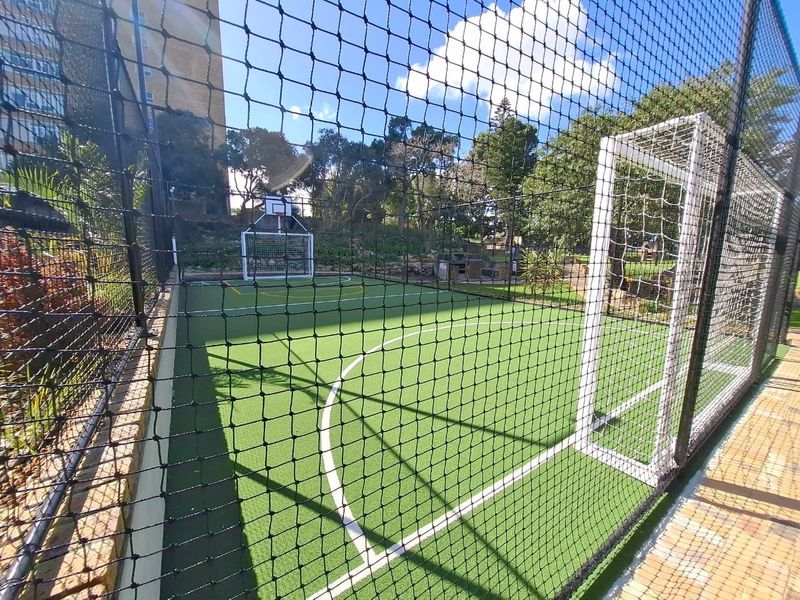 A well-maintained outdoor sports facility featuring artificial turf, soccer goals, a basketball hoop, and secure perimeter netting.