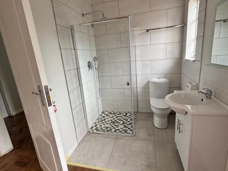 A clean, modern bathroom featuring a glass-enclosed shower, vanity unit with basin, and neutral wall tiling, adjacent to a parquet-floored hallway.