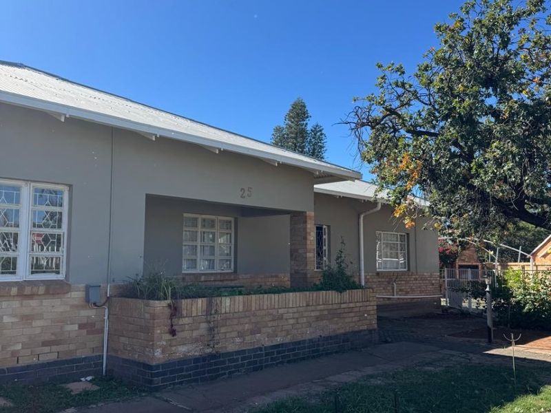 Front elevation of a single-story house featuring a covered porch, brick base, and metal roofing, set within a residential garden area.