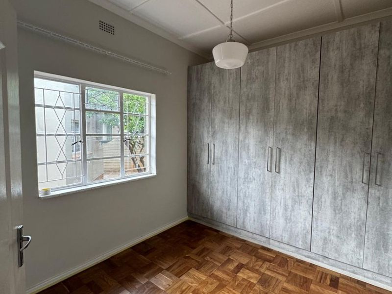 A bright, empty bedroom featuring classic parquet flooring, modern grey built-in cupboards, and a window with security bars.
