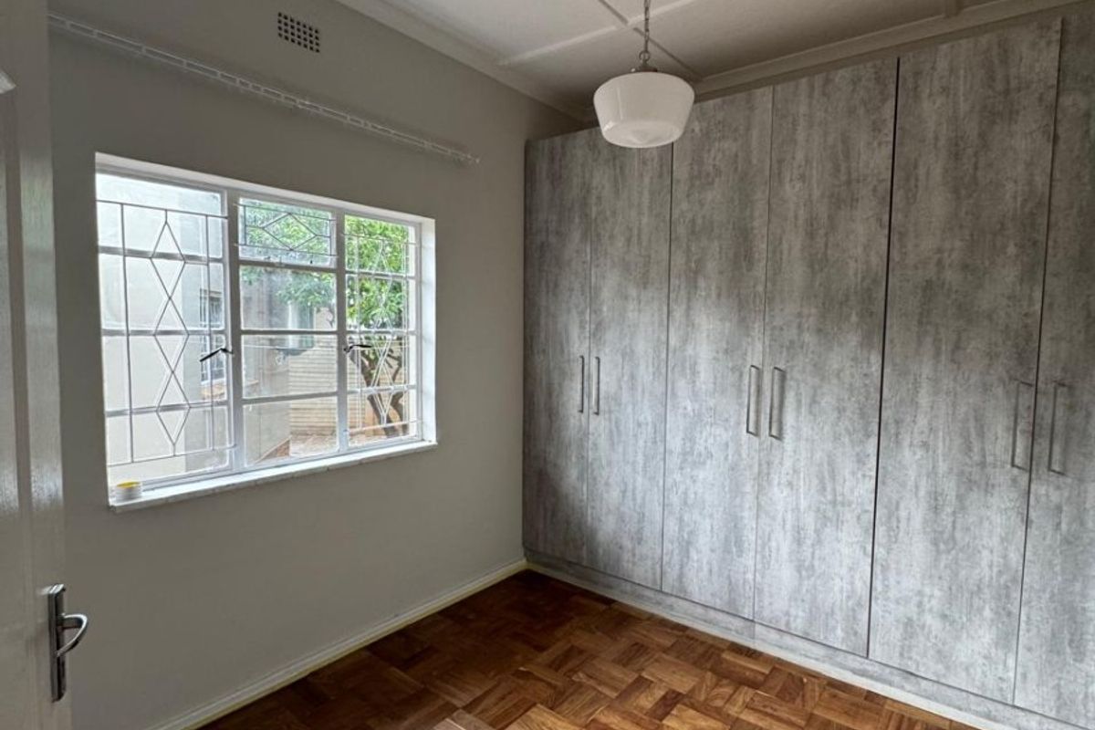 A bright, empty bedroom featuring classic parquet flooring, modern grey built-in cupboards, and a window with security bars.