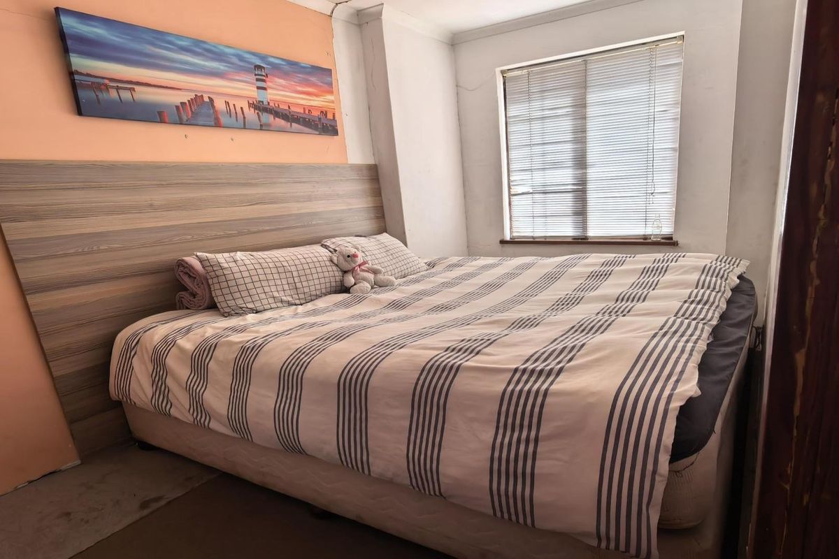 A bedroom featuring a built-in laminate headboard and venetian blinds, showing signs of wear including peeling wall paint and damaged door trim.