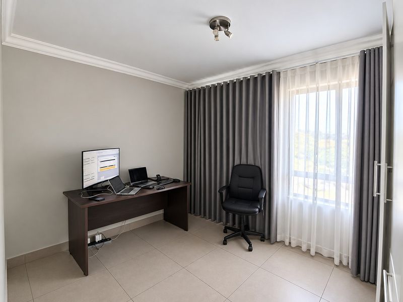 A well-maintained home office featuring neutral tiled flooring, ceiling coving, and floor-to-ceiling curtains providing natural light.