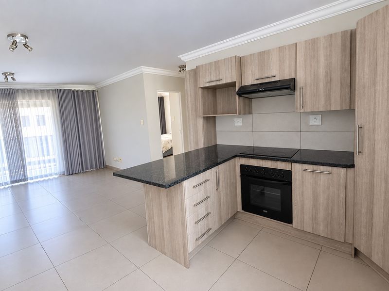 Open-plan kitchen and living area featuring modern wood-finish cabinetry, granite countertops, built-in oven and hob, and neutral tiled flooring throughout.