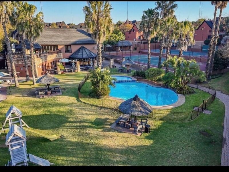 Aerial view of the complex communal area featuring a swimming pool, tennis court, landscaped gardens, paved walkways, and a thatched entertainment area.