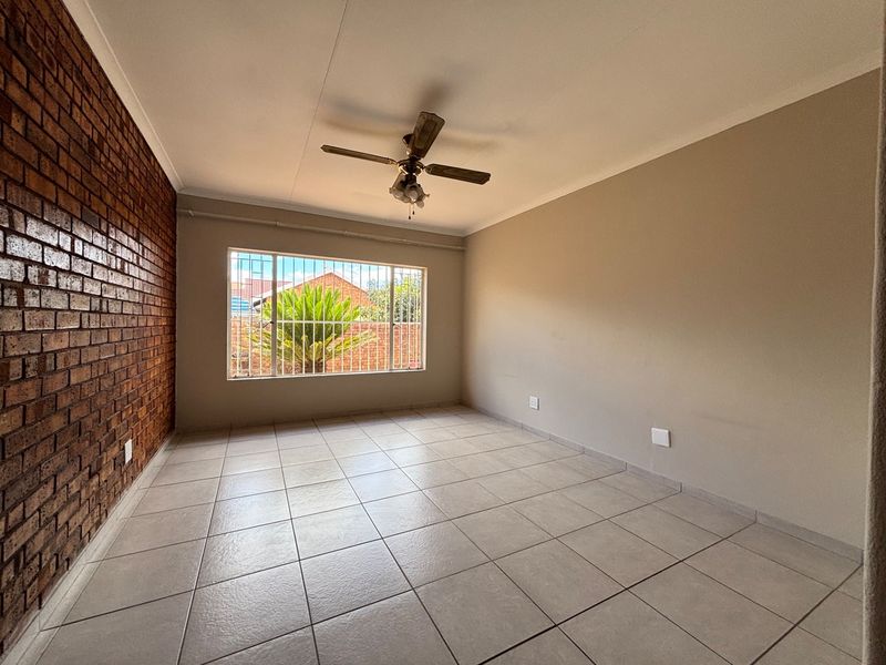 A spacious, empty living room featuring a rustic exposed brick wall, neutral ceramic floor tiles, and a ceiling fan, offering a clean canvas for styling.