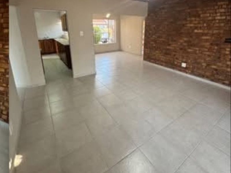 An open-plan living area featuring exposed brick walls, tiled flooring, and a timber ceiling, with an adjacent kitchen visible through the doorway.
