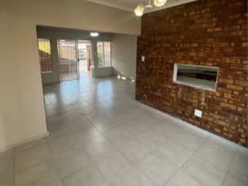 Spacious open-plan living area featuring a distinctive exposed brick wall, neutral tiled flooring, and direct access to an outdoor patio through sliding glass doors.