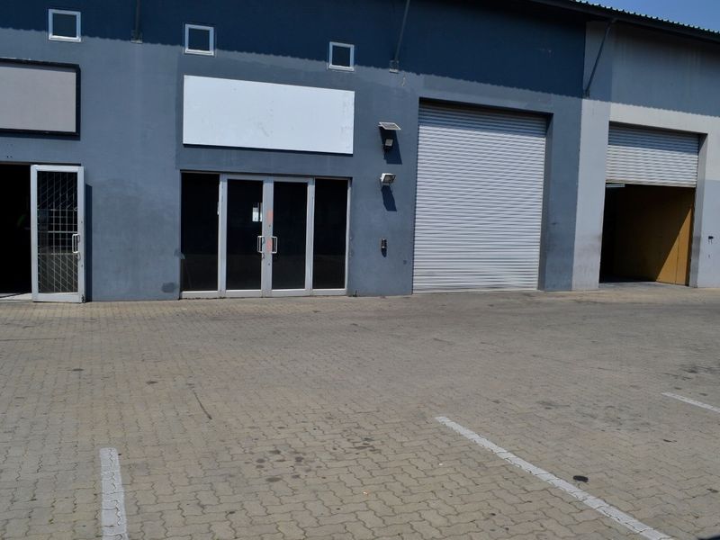 Industrial unit exterior featuring roller shutter doors, glass entrance, and paved frontage, suitable for warehouse or commercial operations.