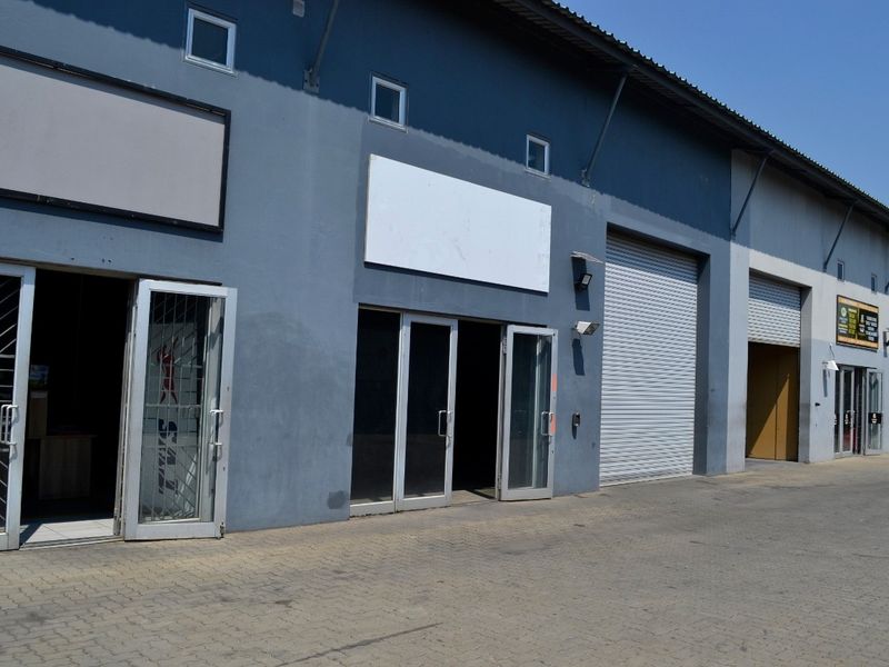 Industrial unit exterior featuring roller shutter doors, glass entrance doors with security gates, and a paved forecourt area.