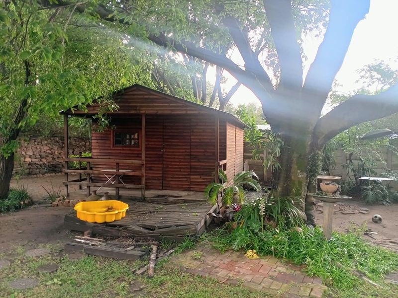A garden setting featuring a wooden cabin with an attached timber deck, set within a mature garden with brick paving and stone boundary walls.