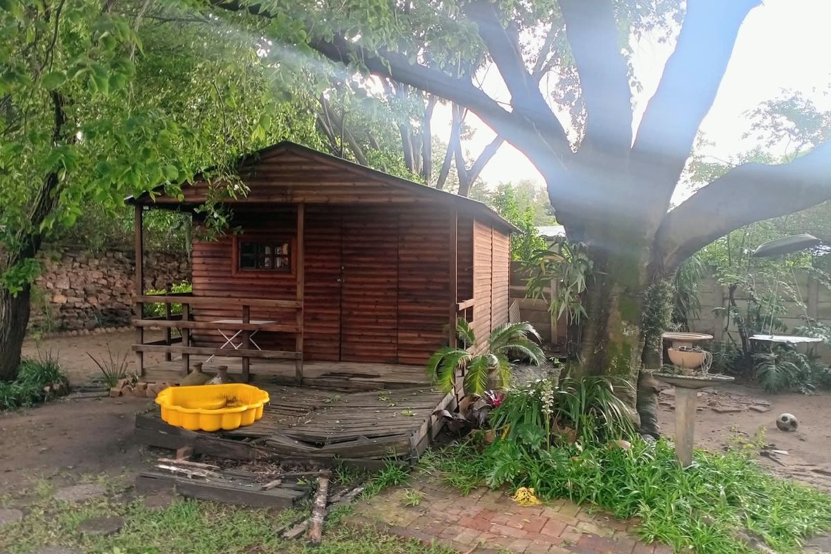 A garden setting featuring a wooden cabin with an attached timber deck, set within a mature garden with brick paving and stone boundary walls.