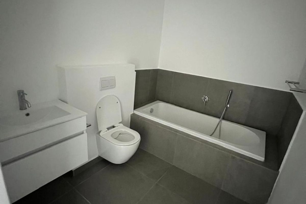 Modern bathroom featuring a wall-hung toilet, built-in bathtub with handheld shower, and a sleek vanity cabinet with basin, finished with neutral floor-to-ceiling tiling.